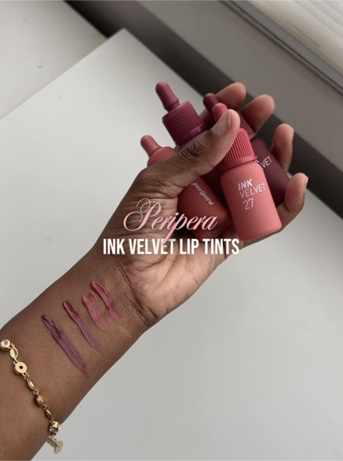 Hand holding three pink lip tint bottles with swatches on wrist, gold bracelet, white background.