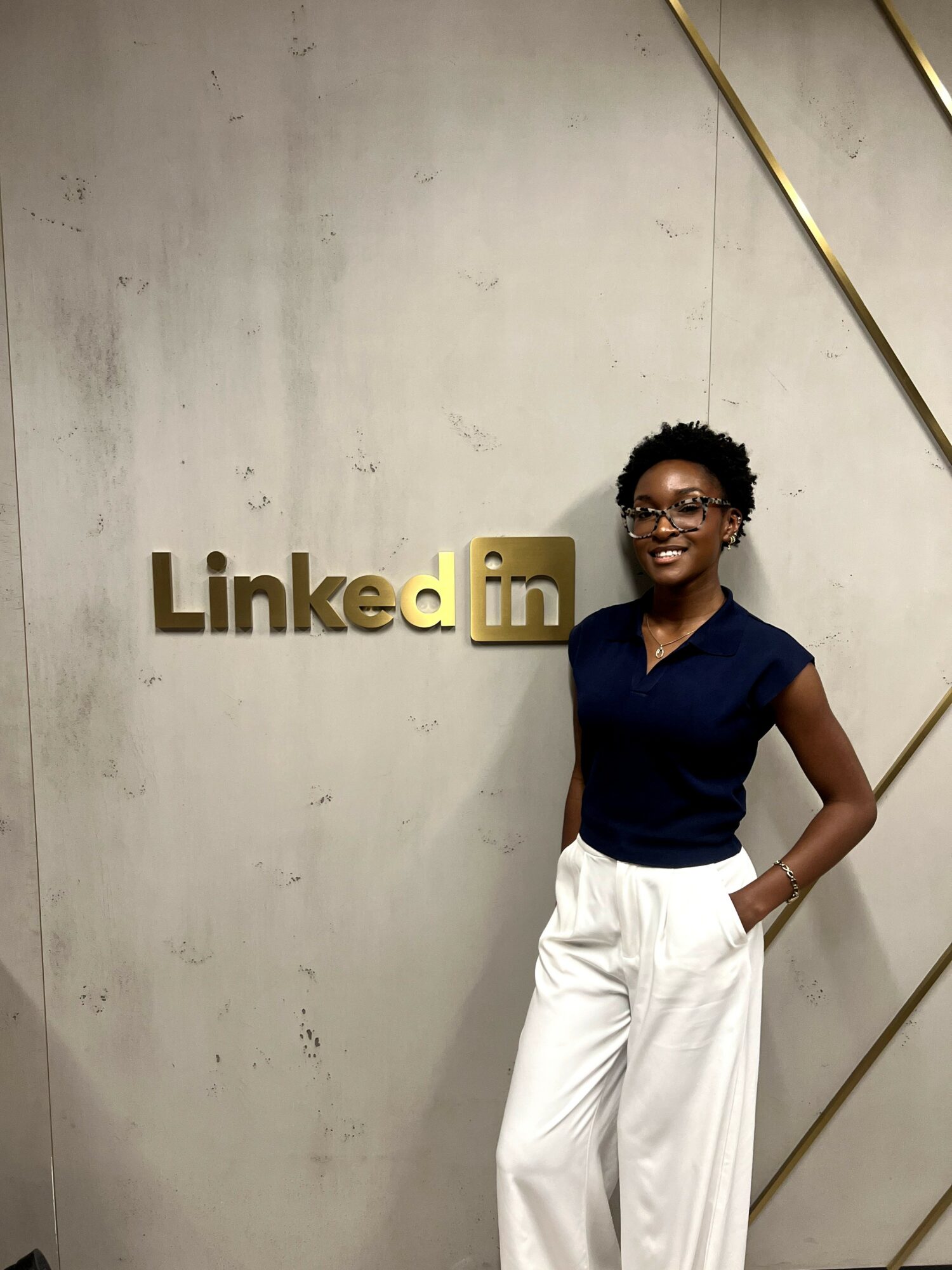 Woman with short curly hair and glasses stands in front of a wall with a LinkedIn sign, smiling, wearing a navy top and white pants.