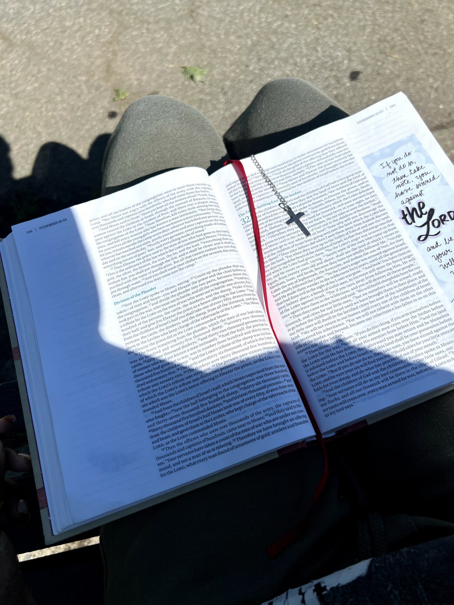 Open book on person's lap with a cross bookmark, shadow cast on pages, sandy ground background.