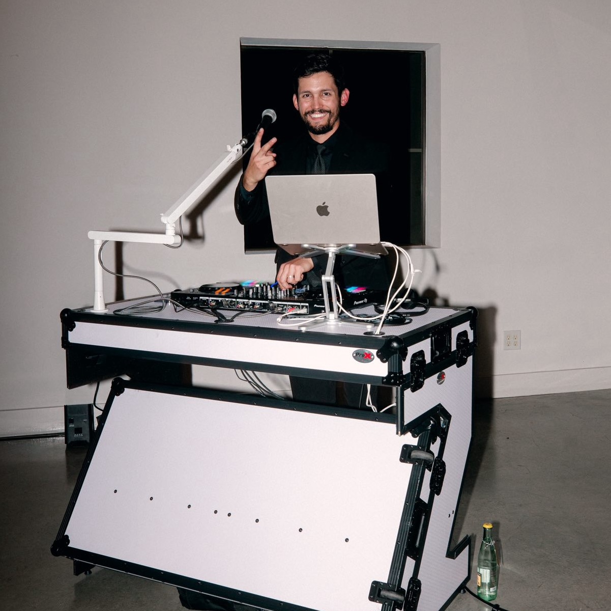Man with a beard smiling and making a peace sign behind a DJ setup with a laptop and equipment, in a room with plain walls.
