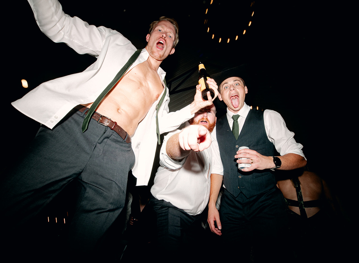 Three men celebrating, one holding a beer bottle, smiling and raising their arms, in a dark setting with string lights.