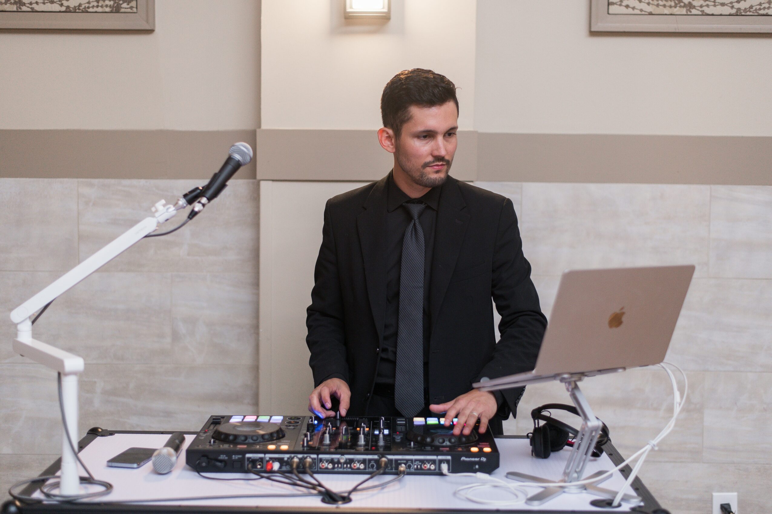 Man in black suit and tie standing behind DJ equipment with laptop and microphone, in a room with light-colored walls.