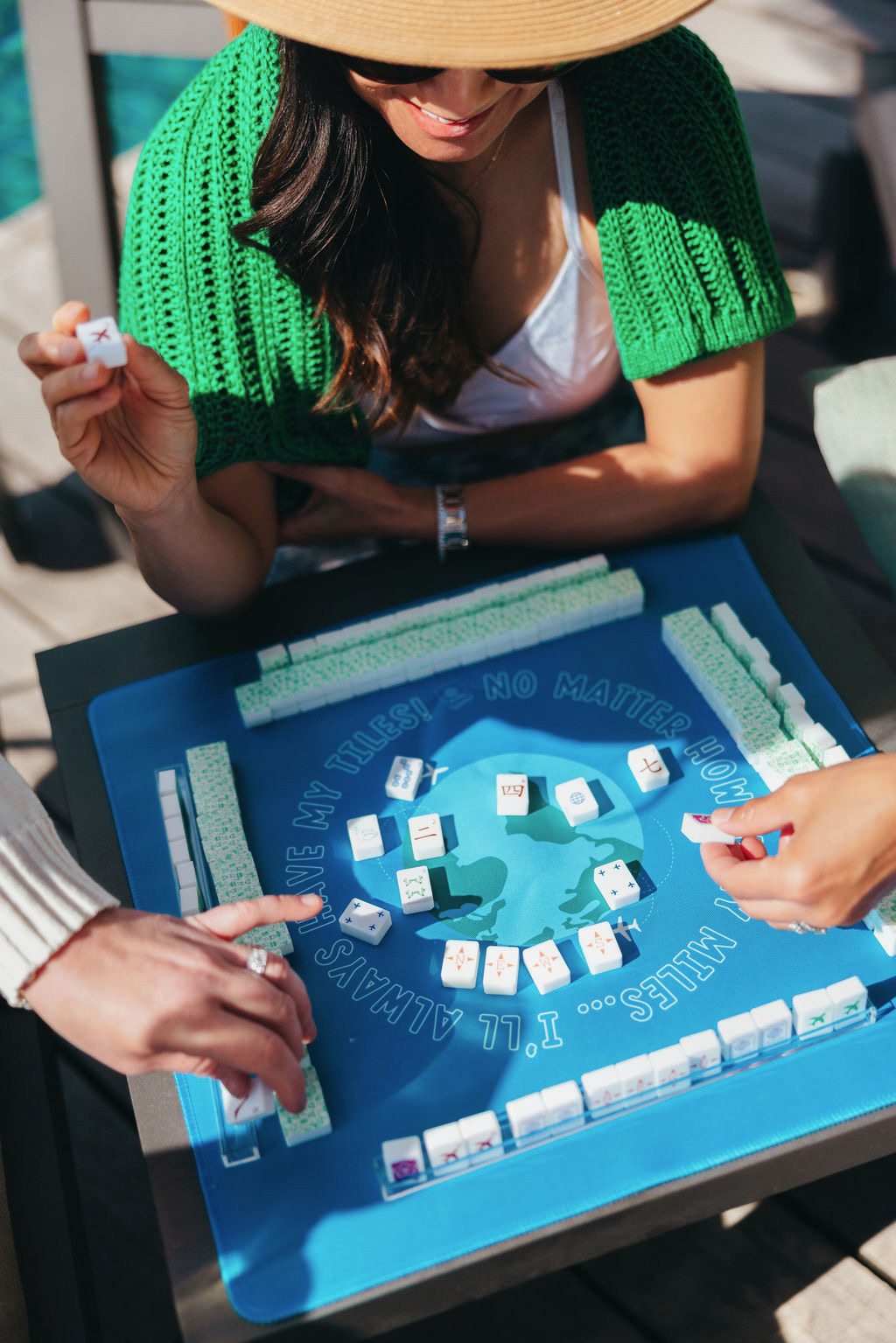 People playing a game with tiles and dice on a blue table outdoors, with a woman wearing a green sweater and hat.