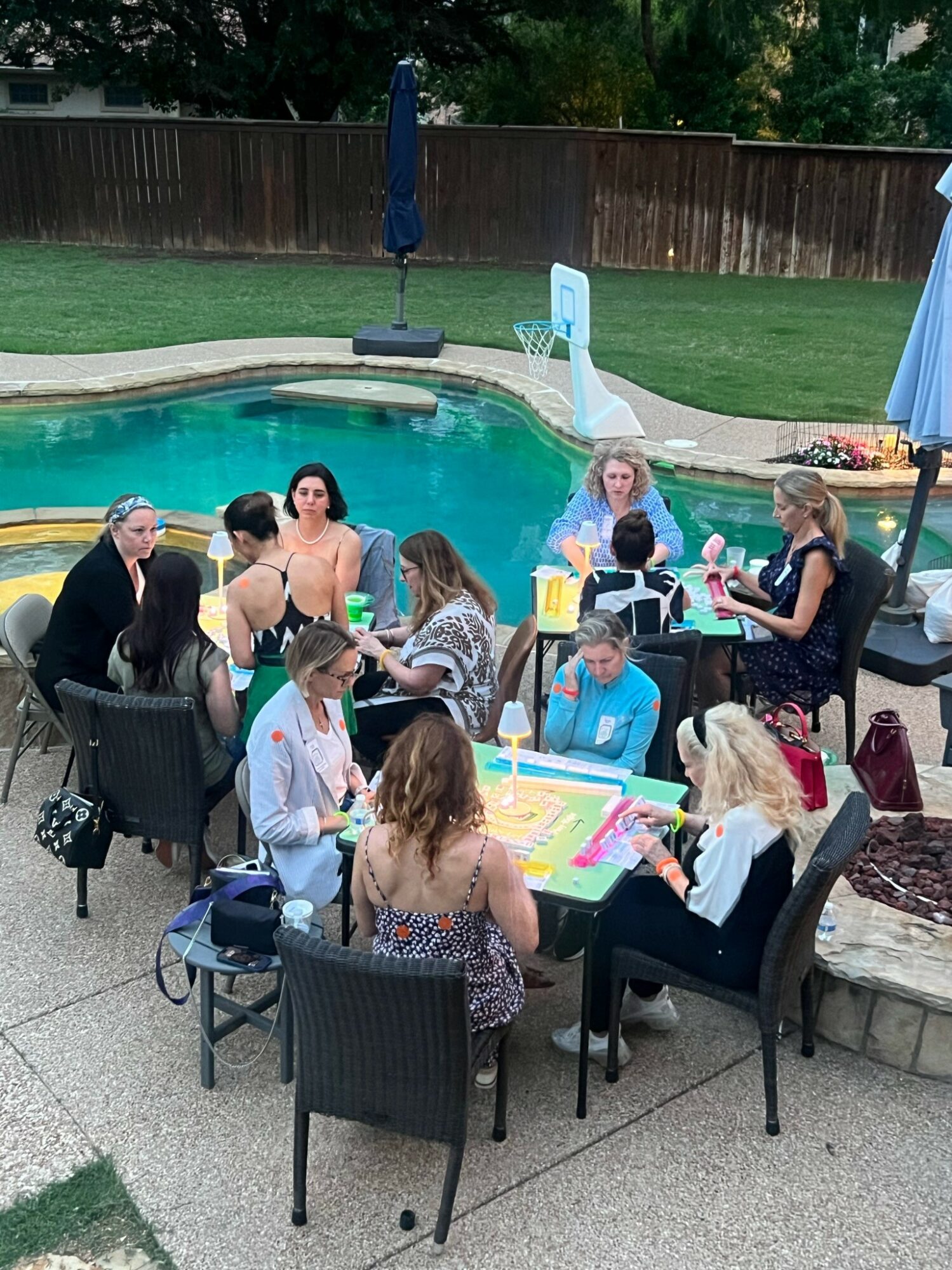 Group of people sitting around a table near a swimming pool outdoors, some with drinks and snacks, in a backyard setting.