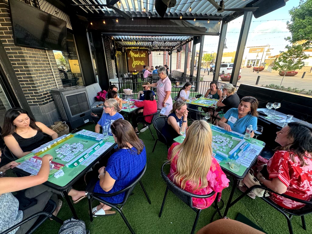 People playing Mahjong at outdoor tables under a covered patio, with some standing and others seated, in a lively setting.