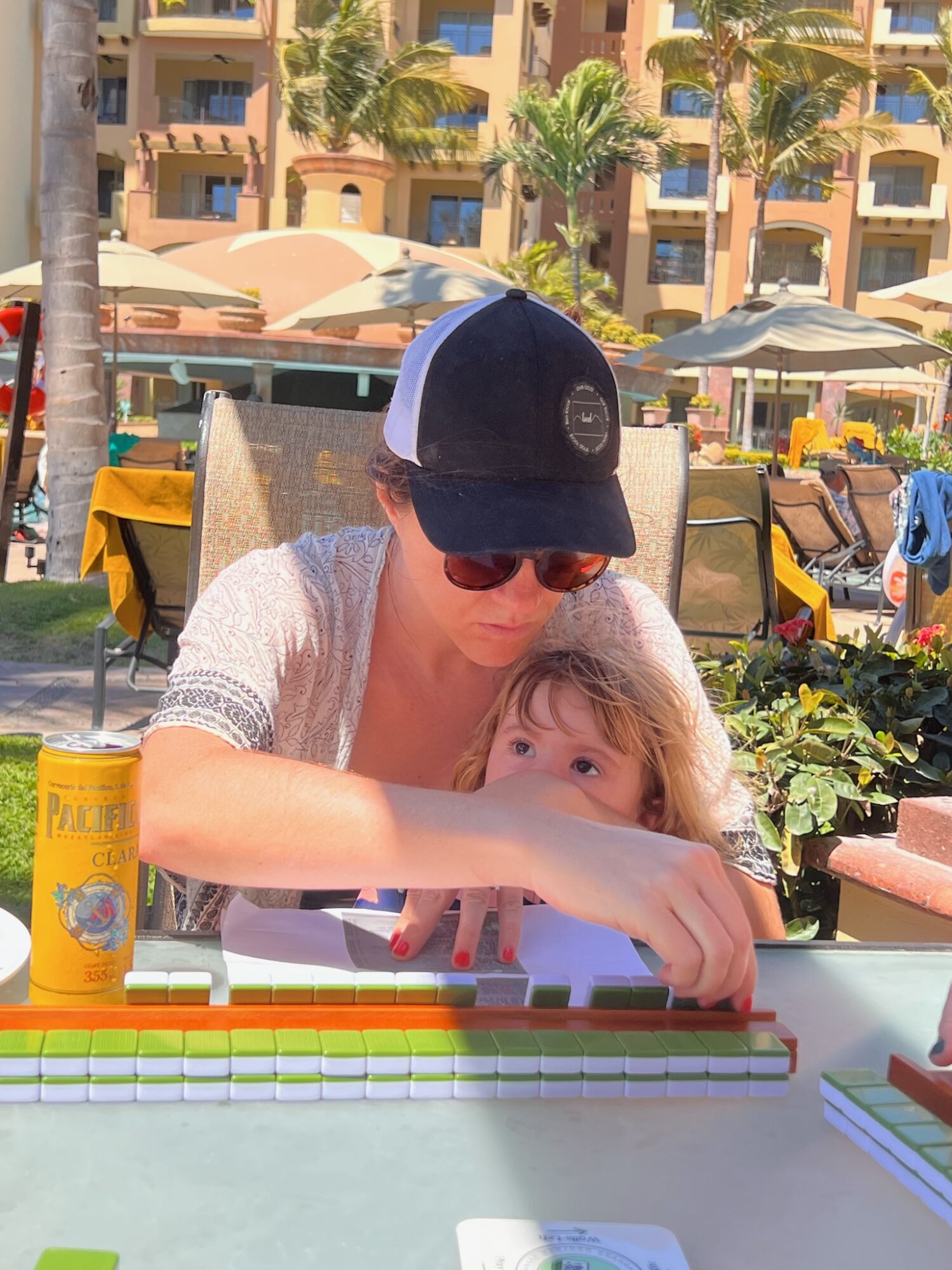 Woman wearing sunglasses and a cap playing a game with a child at an outdoor table, sunny day with palm trees and umbrellas.