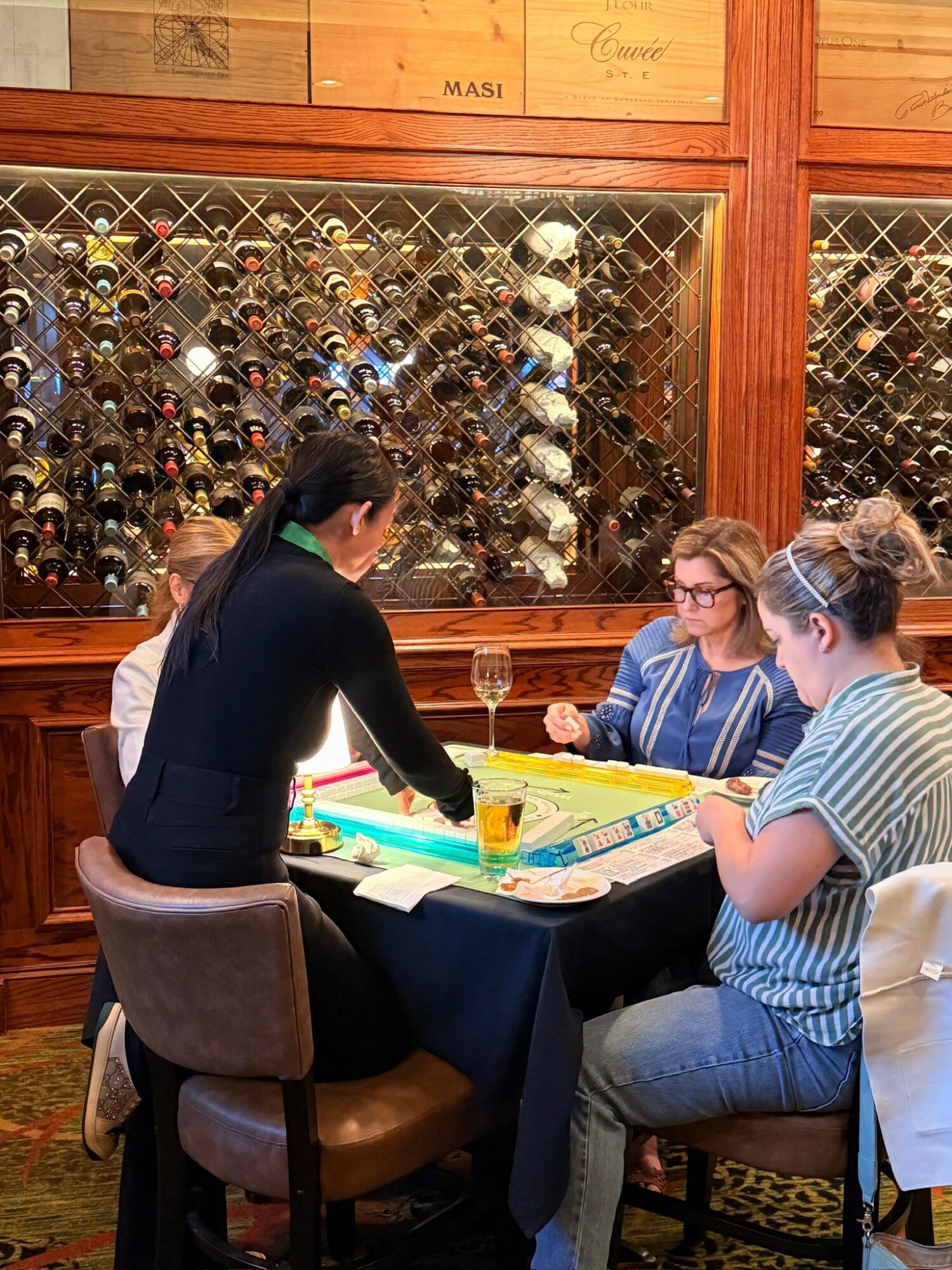 Four people sit around a table playing a game, with a wine glass and game pieces visible, in a room with a wine display behind them.