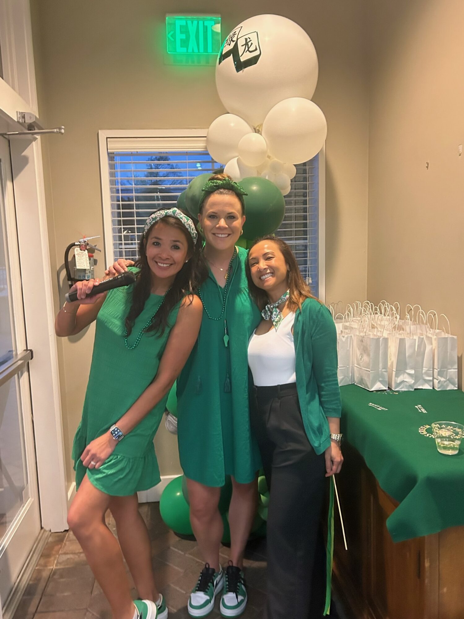 Three women standing together indoors, smiling, with green and white balloons behind them, near a table with drinks and a window.