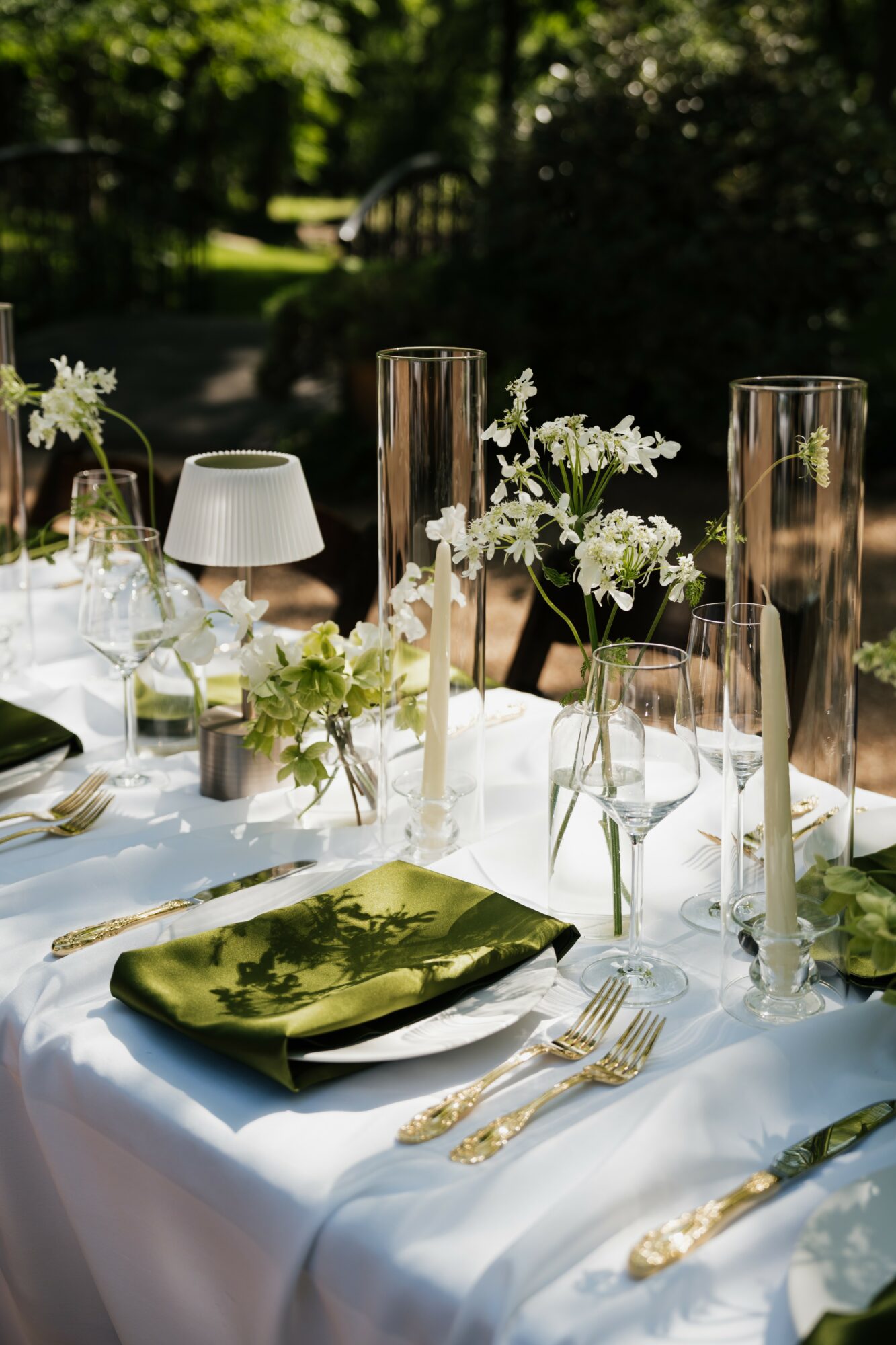 Table set outdoors with white flowers, glass vases, candles, and gold cutlery, illuminated by sunlight.