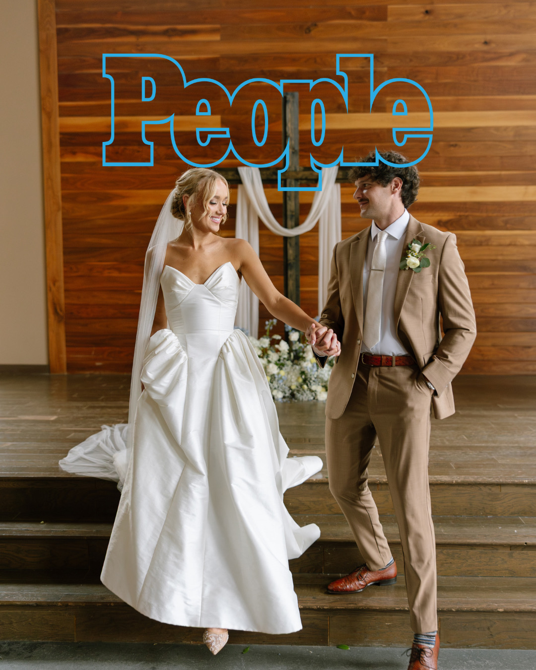Bride and groom holding hands on stage with a large blue 'People' sign behind them, wedding attire, smiling, wooden background.