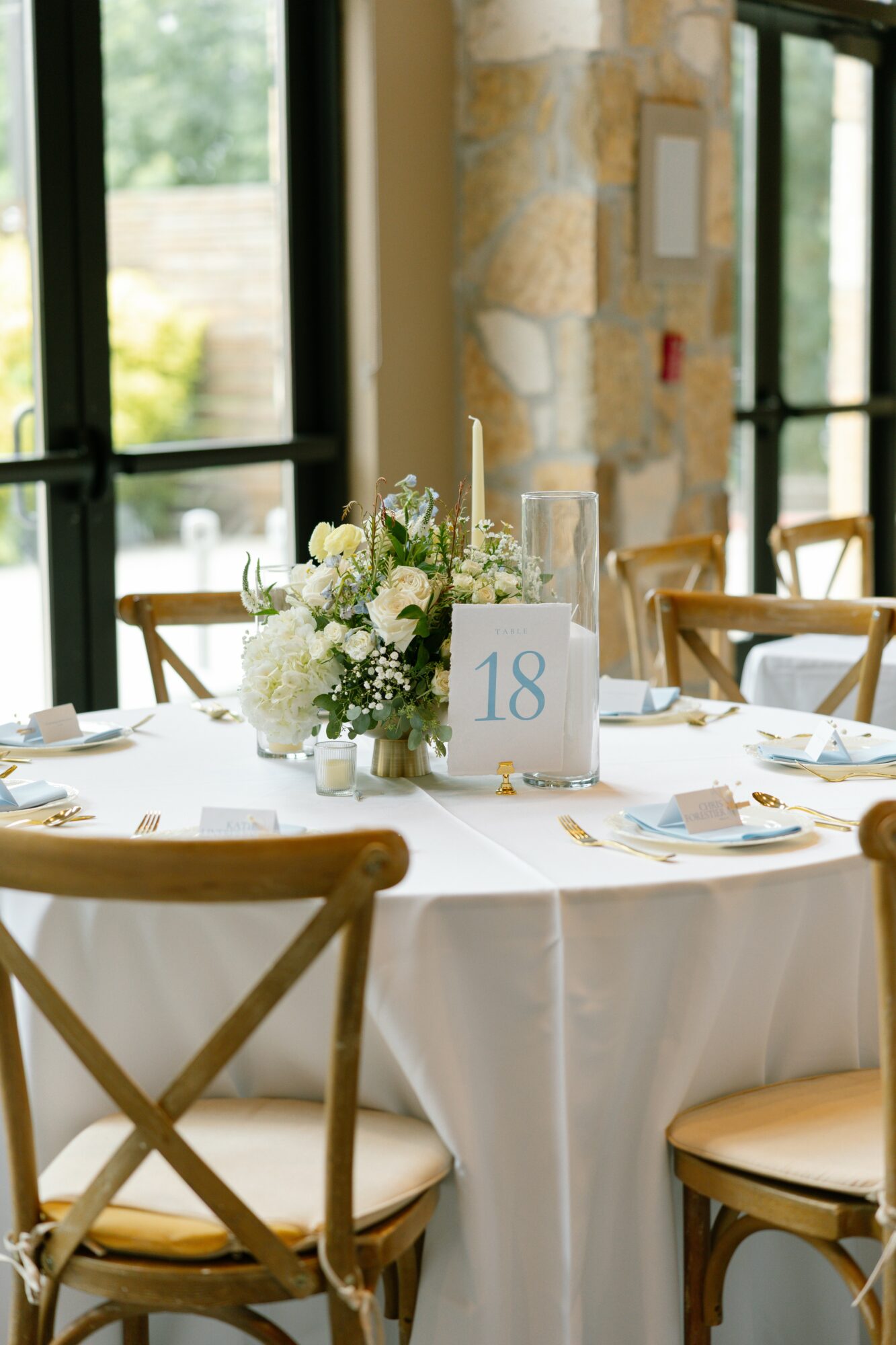 Round table with white tablecloth, floral centerpiece, candles, and table number 18 in a bright room with large windows.