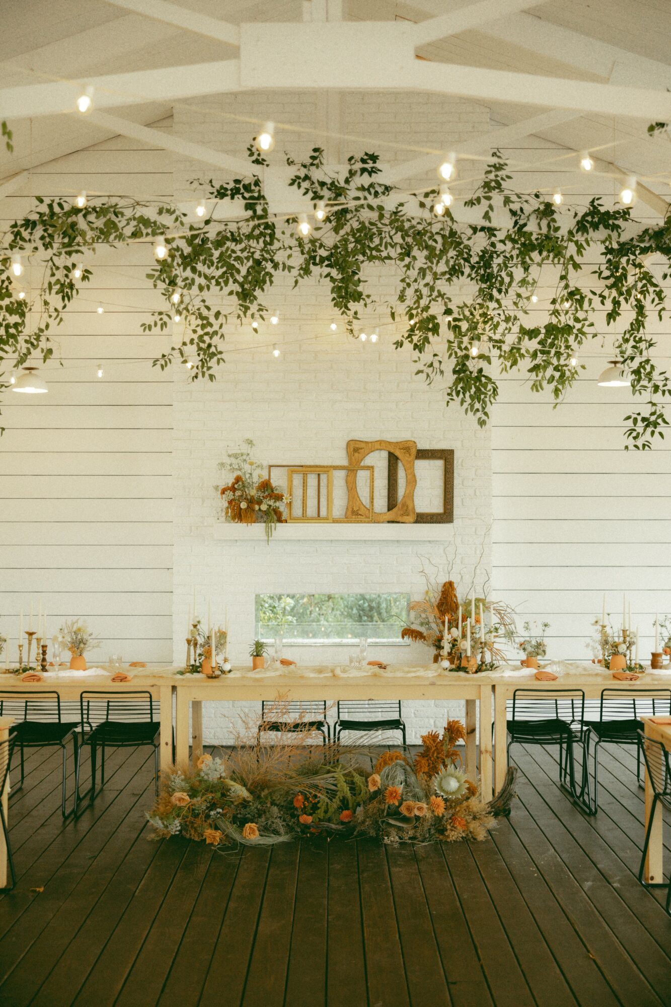 Decorated table with chairs, greenery hanging above, and a white wall with shelves and window, set for an event.
