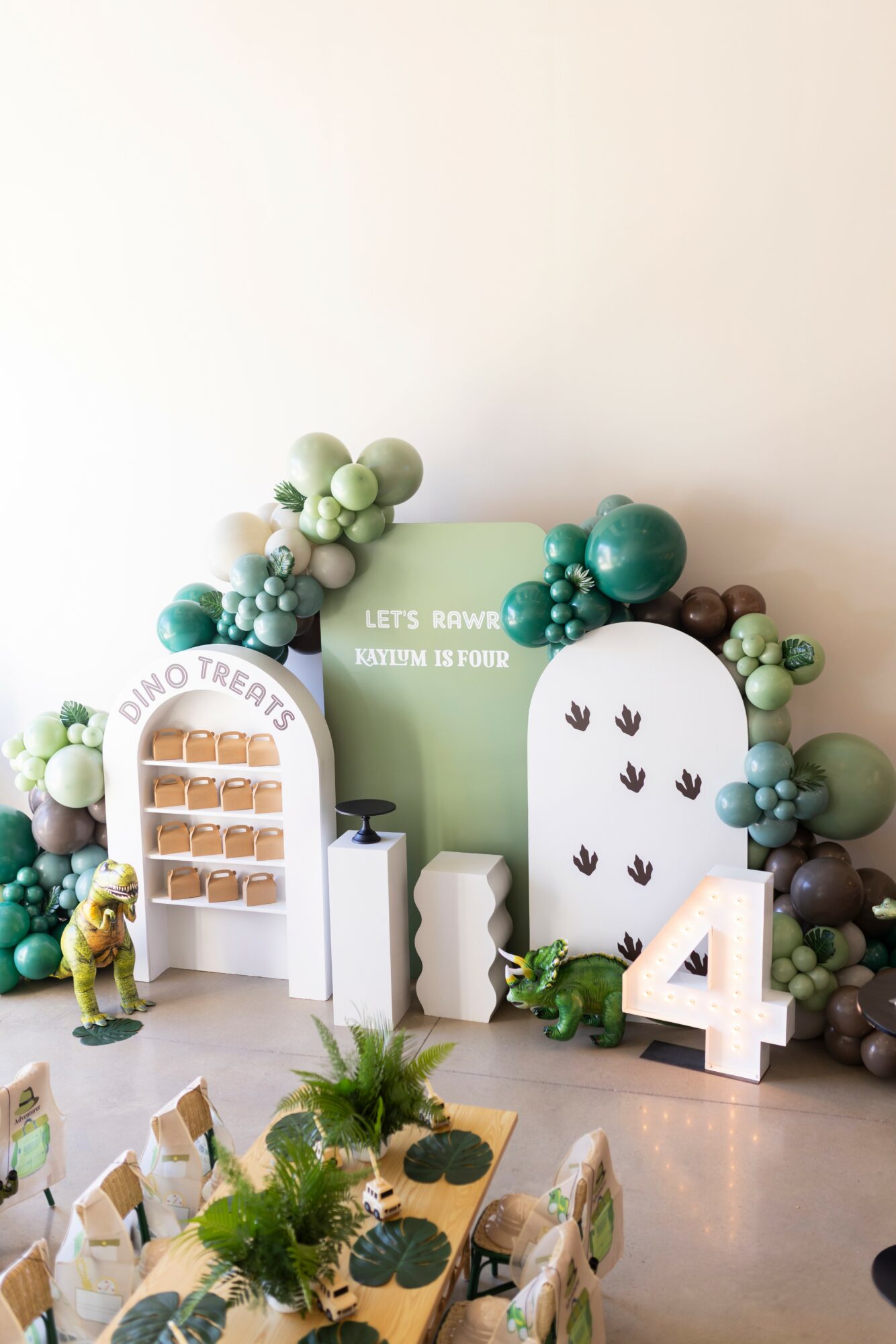 Decorative birthday setup with trees, a large number 4, and small animal figures, on a table with chairs, against a white wall.