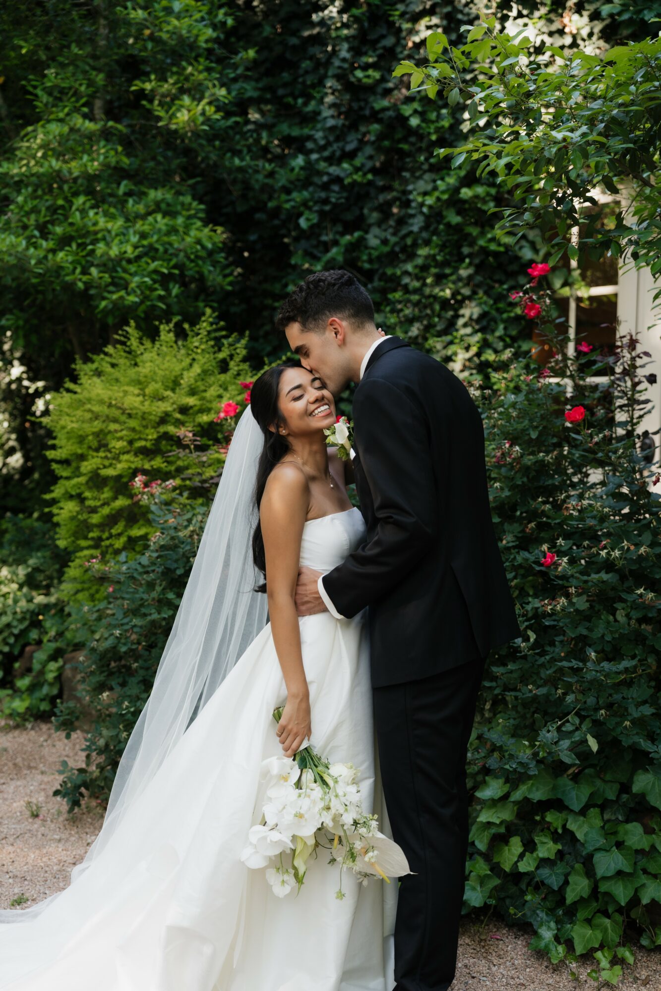 Bride and groom standing close in a garden, surrounded by greenery and flowers, with the bride holding a bouquet.