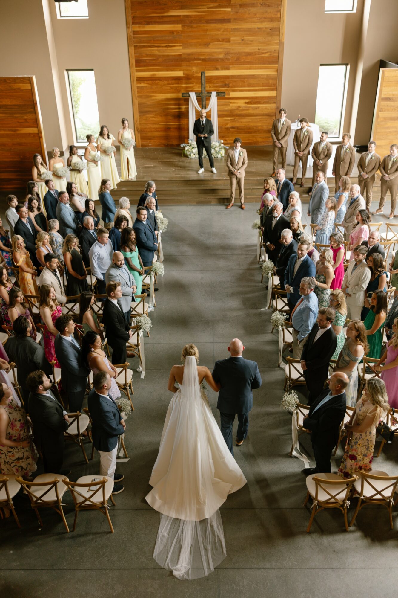 Bride and groom walking down aisle with guests seated on both sides in a church, wooden wall backdrop, sunlight through windows.