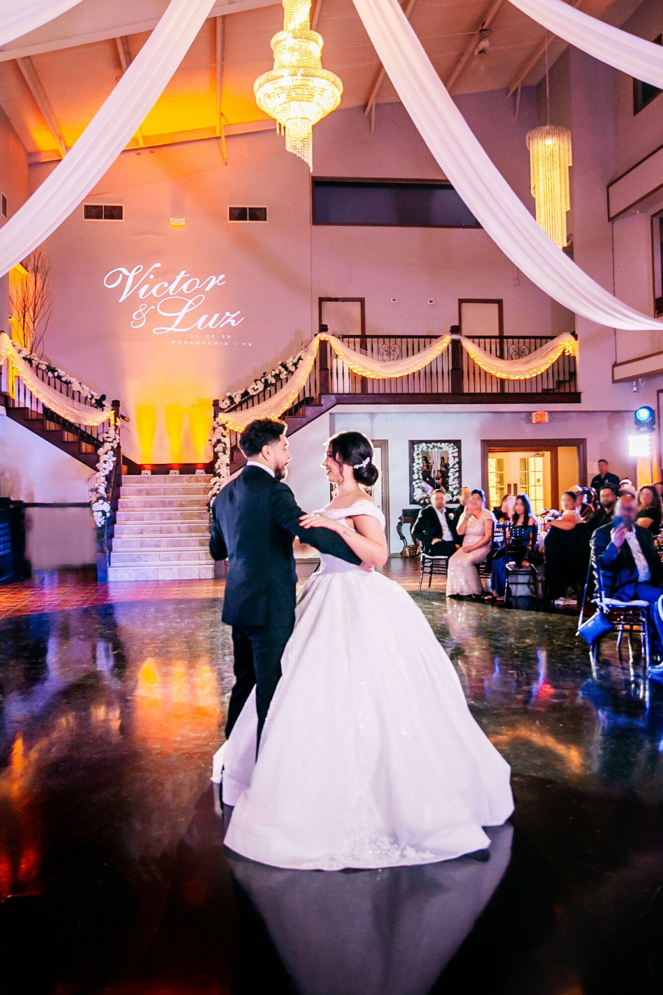 Couple dancing in a decorated ballroom with draped fabric and chandeliers, guests seated around the dance floor, wedding projection on wall.