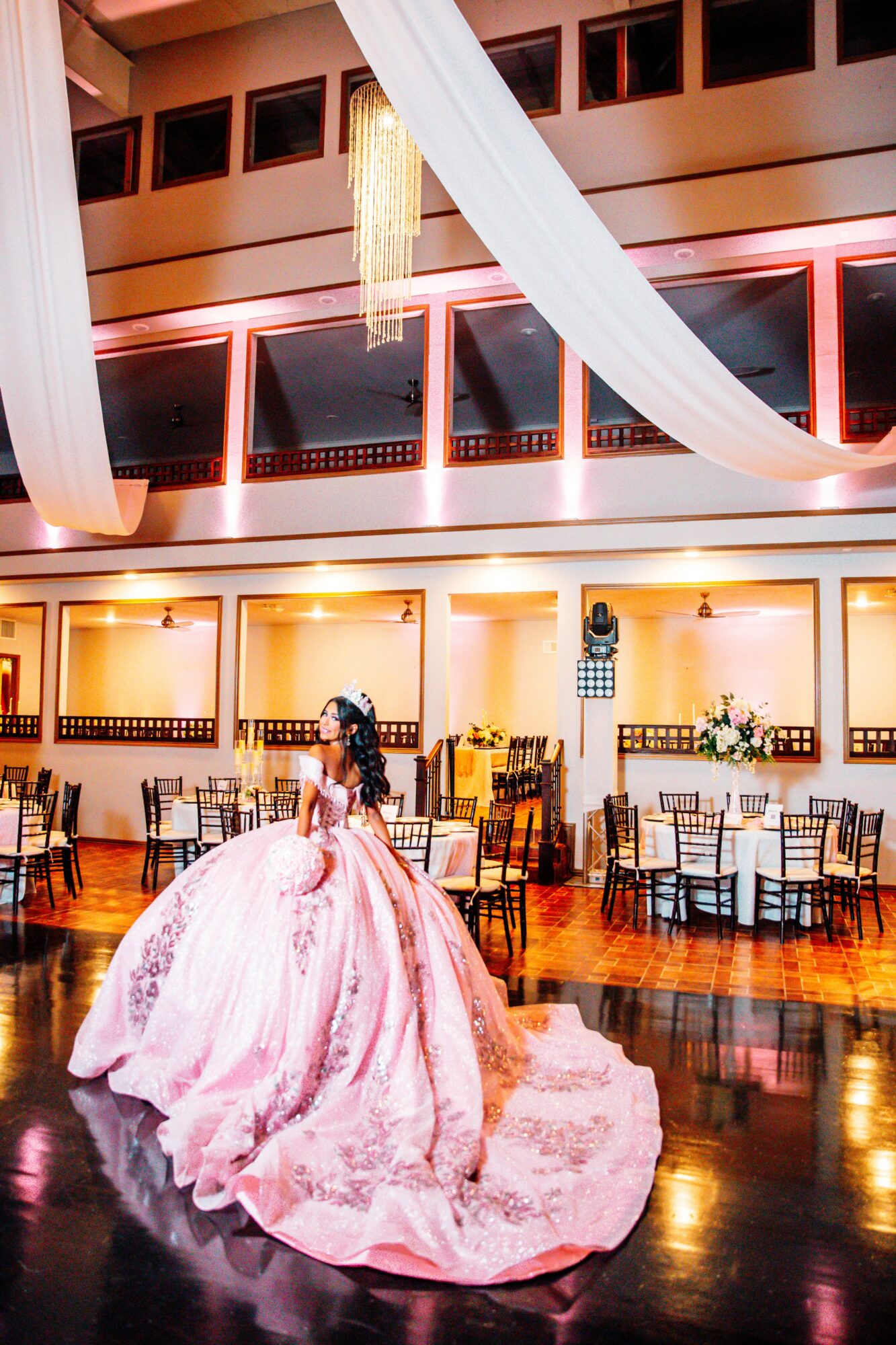 A woman in a pink ball gown stands in a decorated event hall with tables and drapes, illuminated by warm lighting.