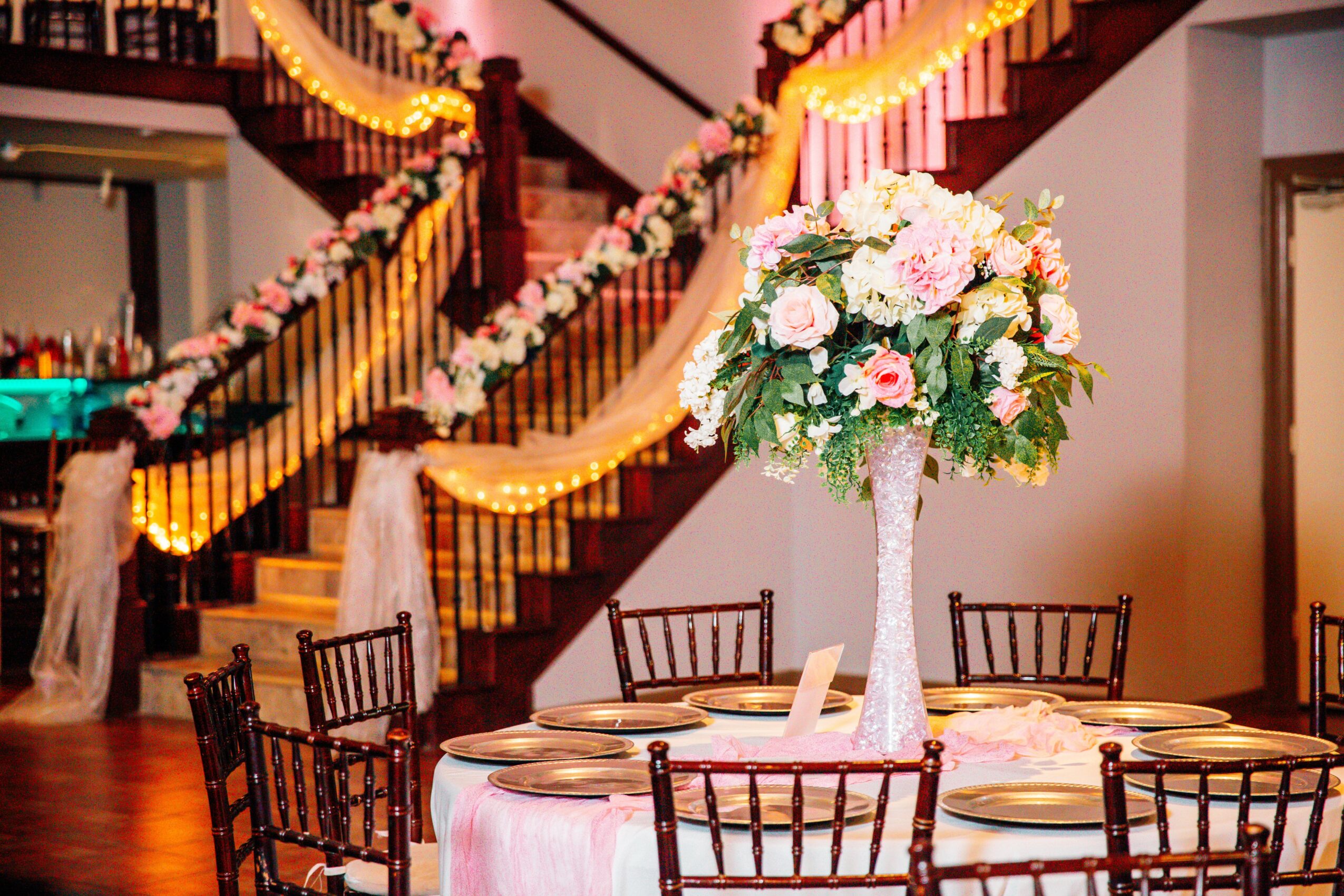 Round table with a tall floral centerpiece, surrounded by chairs, decorated staircase with flowers and drapes in the background.