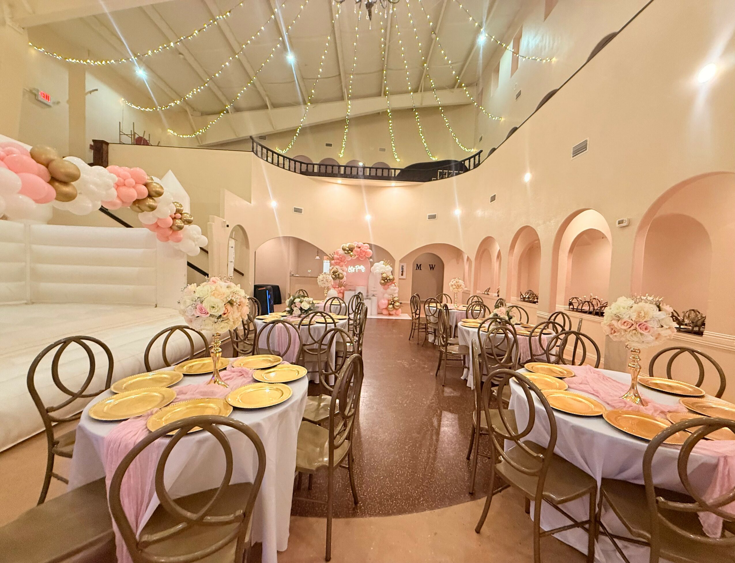 Elegant banquet hall decorated with pink and white flowers, round tables with pink tablecloths, and string lights on the ceiling.