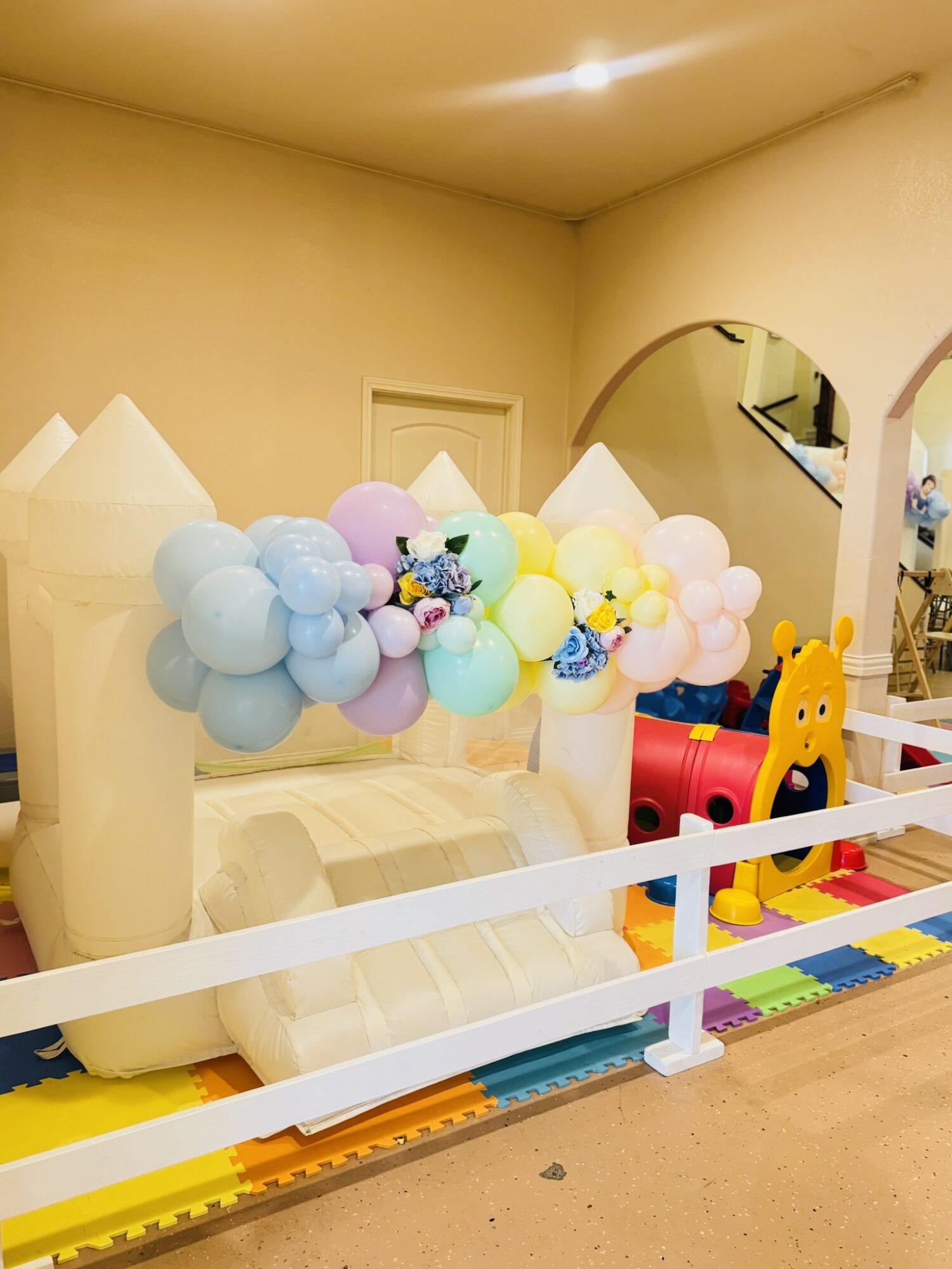 Indoor play area with colorful balloon decorations, a slide, and a cartoon character figure, enclosed by a white barrier.