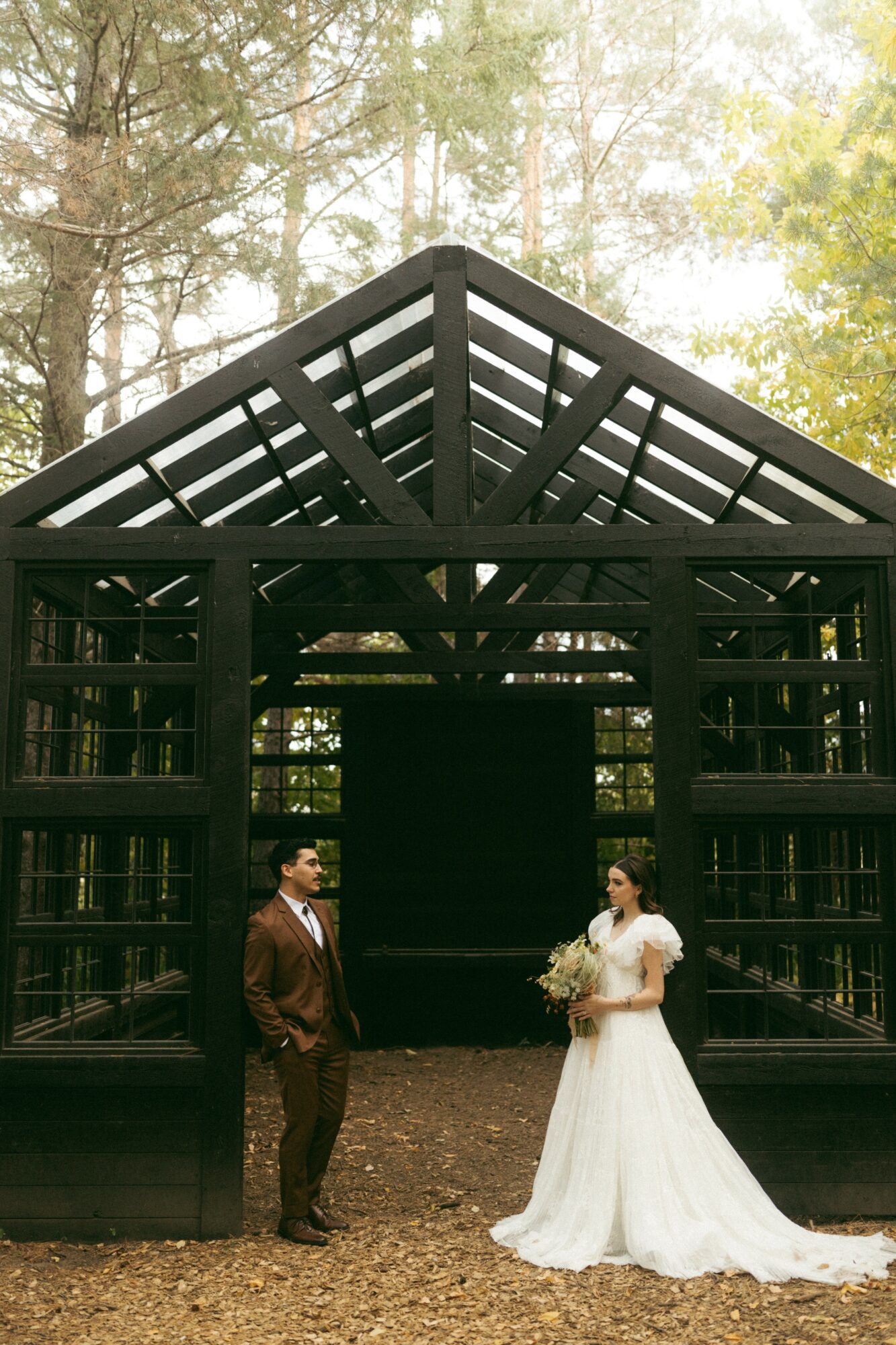 A bride in a white wedding dress holding a bouquet and a groom in a suit stand outside a black wooden structure in a forest setting.