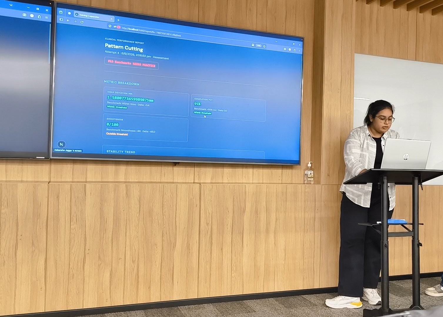 Person standing at a podium in front of a large screen displaying a blue interface, in a room with wooden walls.