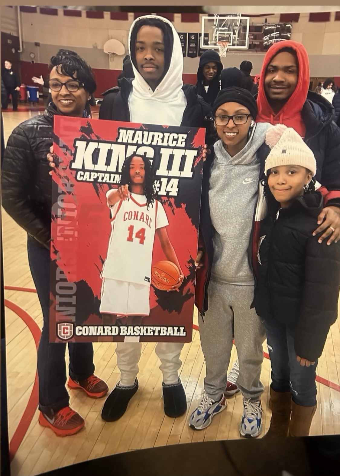 Group of six people, five women and one man, standing in a gymnasium, holding a basketball-themed poster, smiling.