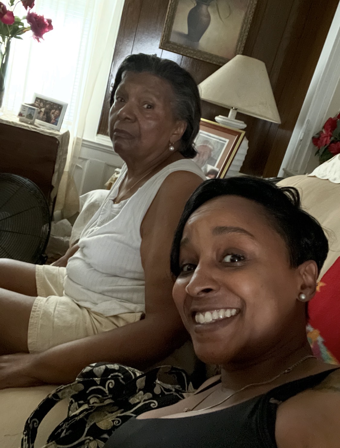 Two women sitting indoors, one smiling at the camera, the other looking away, with a lamp and window in the background.