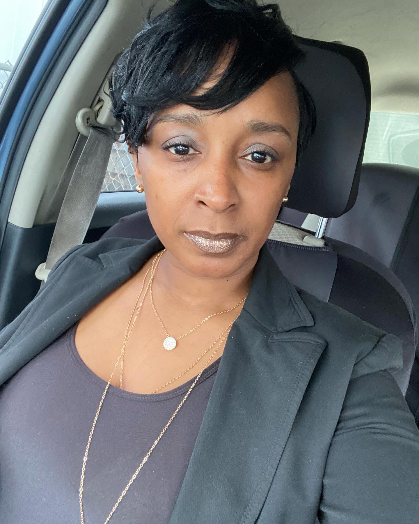 Woman with short black hair wearing a black blazer and gold necklaces, sitting in a car, looking at the camera.