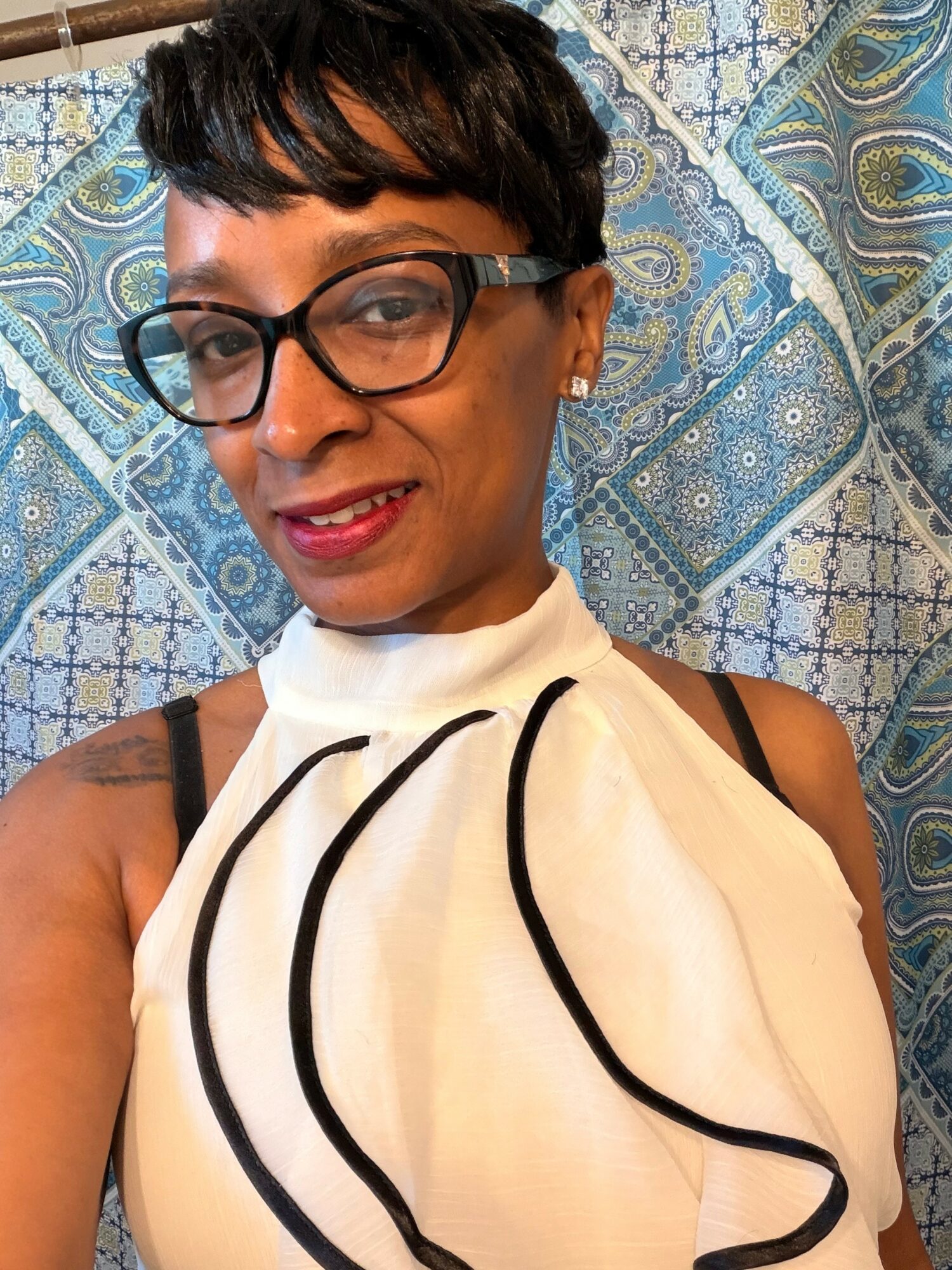 Woman with short hair, glasses, and a white top with black lines, smiling, in front of a patterned blue background.