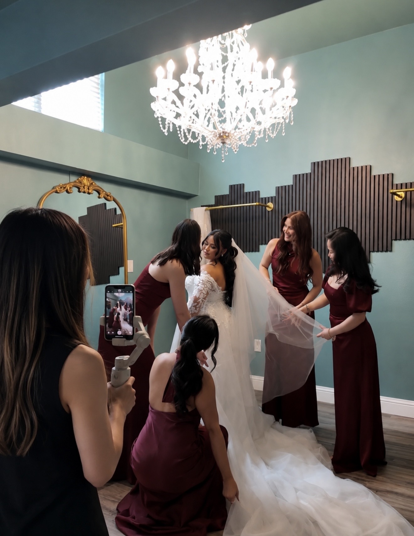 Group of women, including a bride in a wedding dress, gathered in a decorated room with a chandelier and mirror, taking photos.