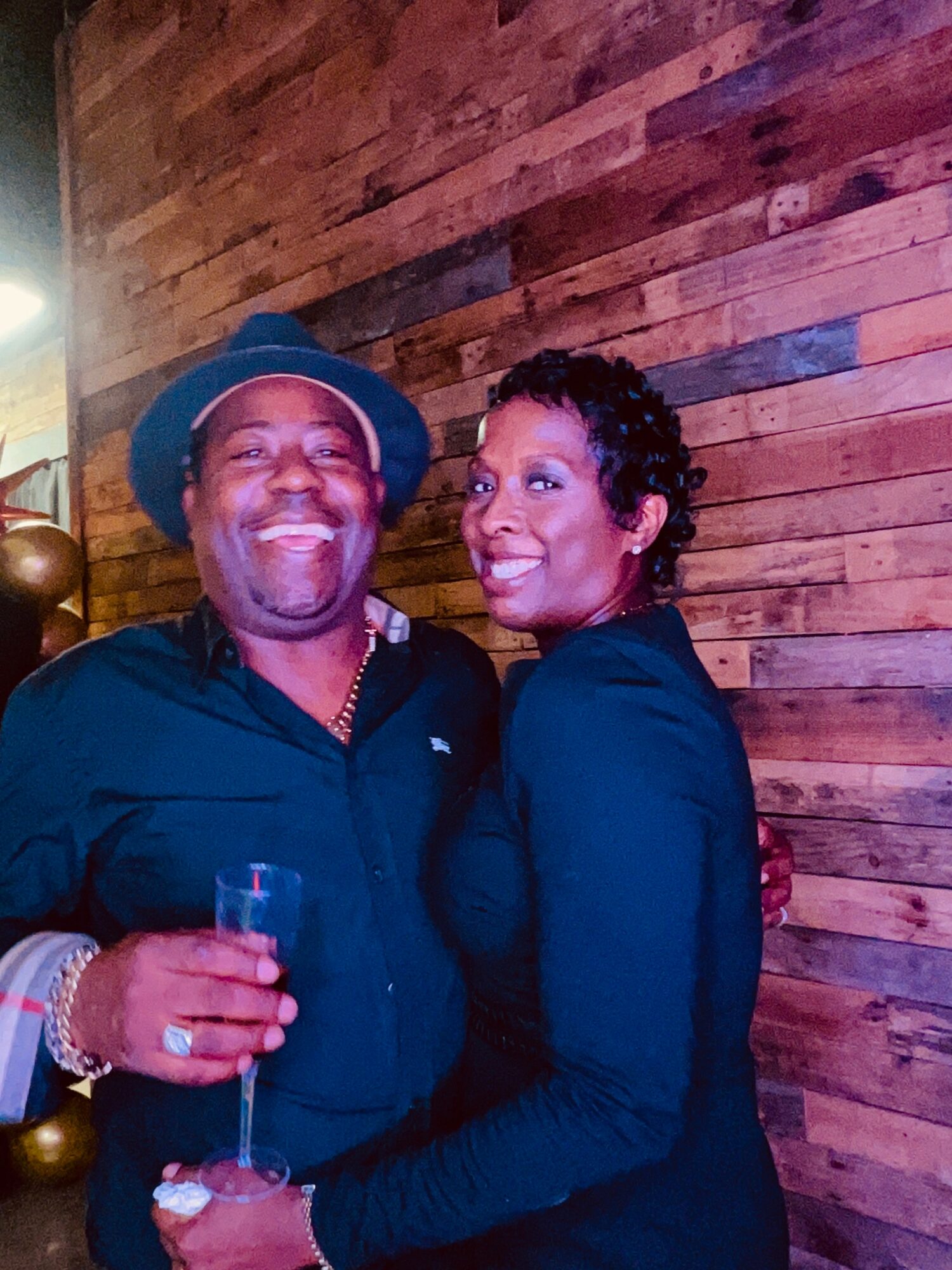 Two people smiling and posing together indoors, one holding a glass, against a wooden wall background.
