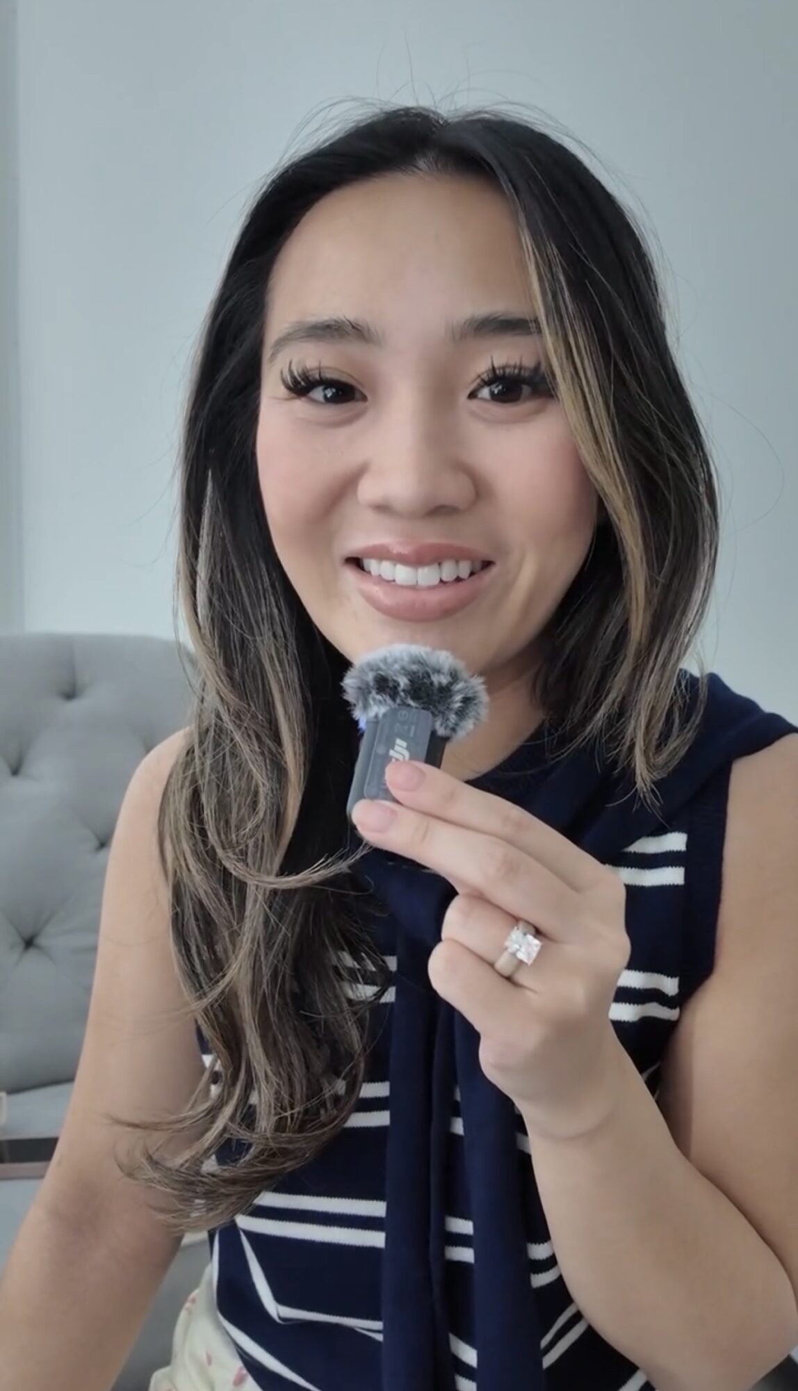 Woman with long hair holding a small object, smiling, in a room with a light-colored wall and sofa behind her.