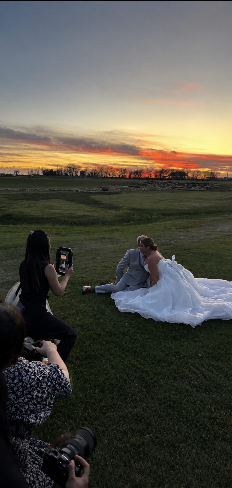 People taking photos of a bride in a white wedding dress on a grassy field during sunset.