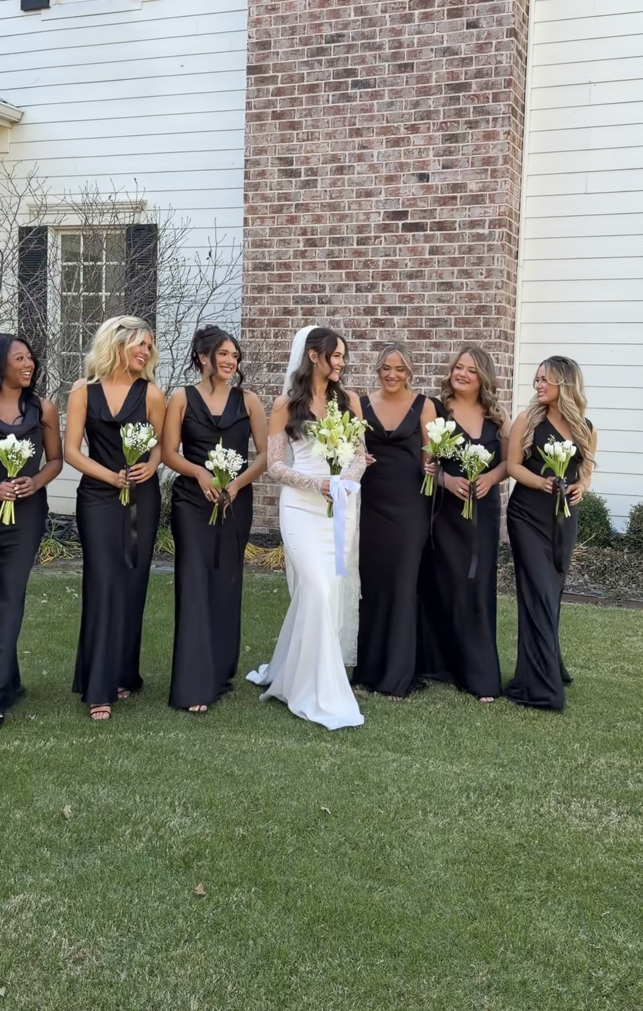 Bride in white dress with veil and six bridesmaids in black dresses holding bouquets, standing on grass in front of a brick and white building.