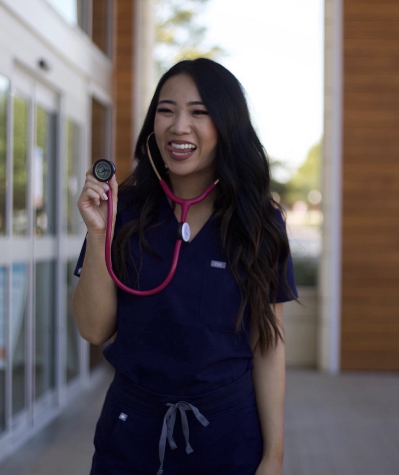 Smiling woman in medical scrubs holding a stethoscope, standing outdoors near a building with glass windows.