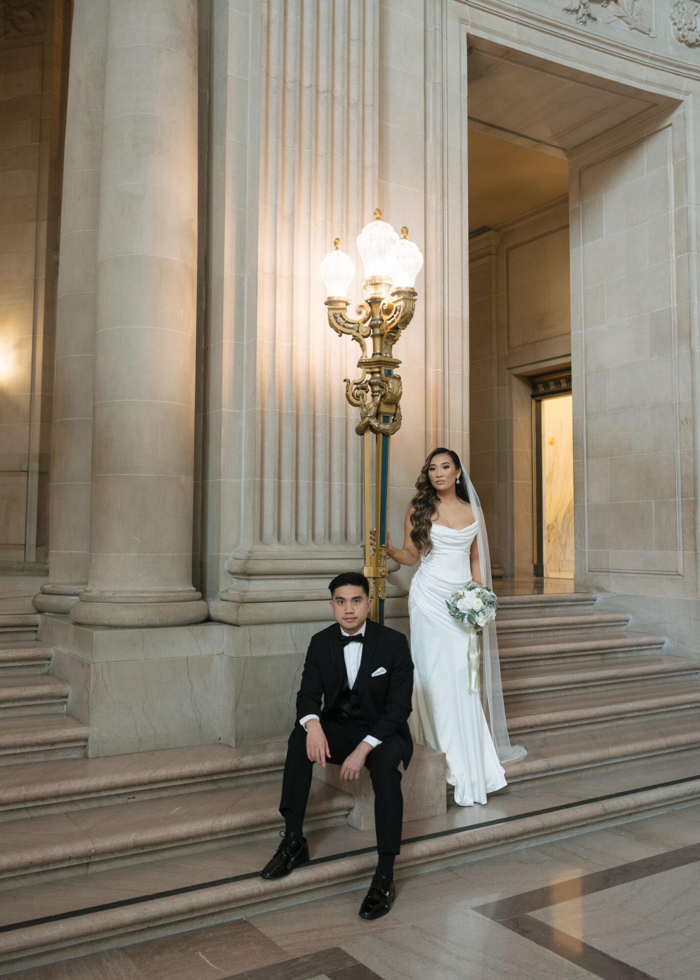A bride and groom pose on stairs in a grand, ornate building with tall columns and a decorative lamp post.