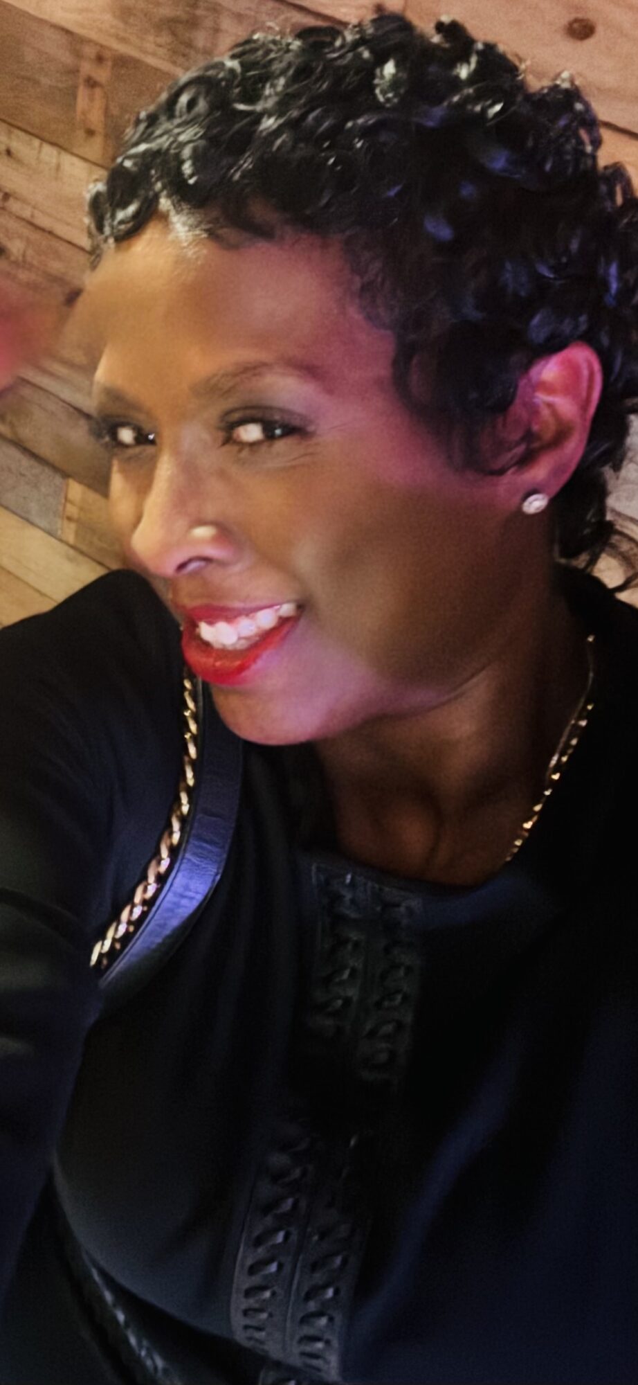 Close-up of a woman with short curly hair, smiling, wearing a black top and a gold chain, wooden background