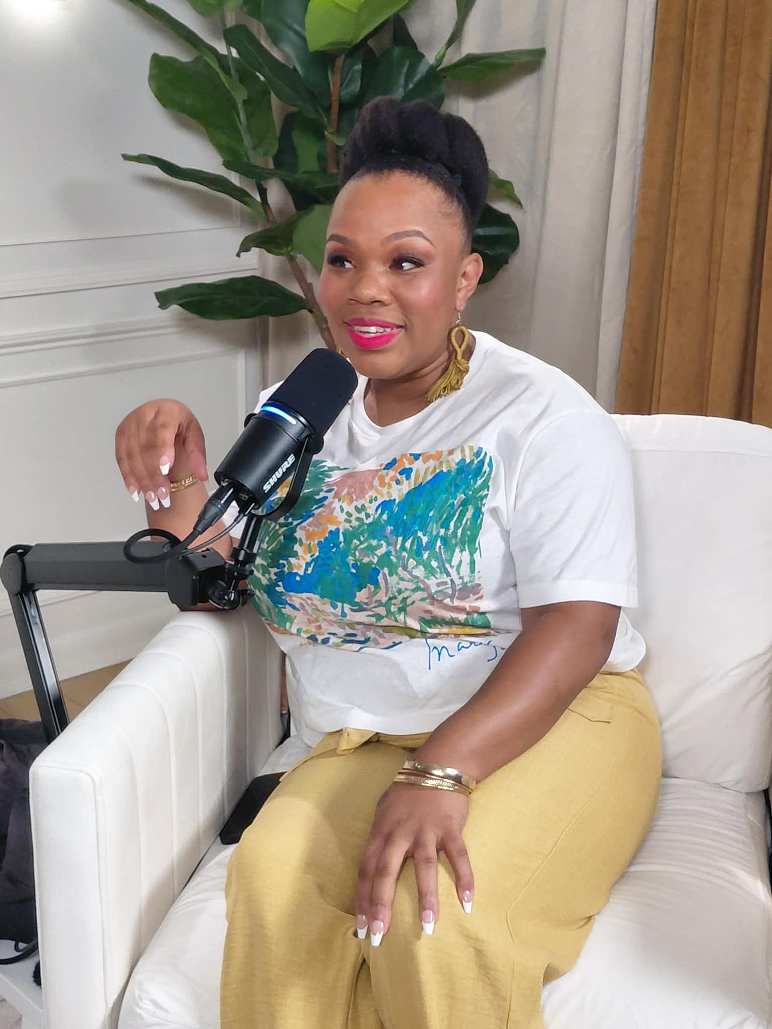 Woman with short hair, wearing a white T-shirt with a colorful design, sitting on a white couch, speaking into a microphone.