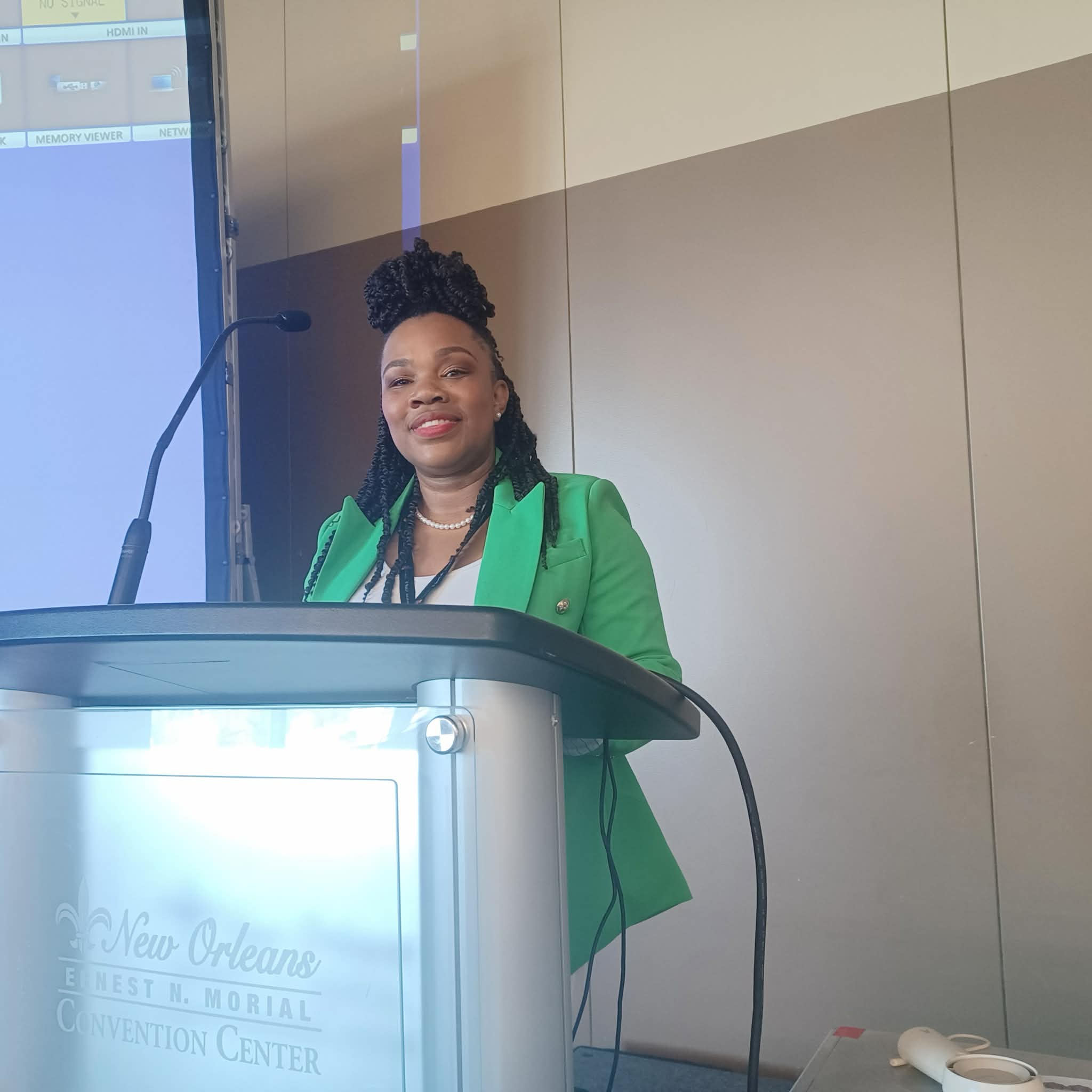 Woman standing at a podium, smiling, wearing a green blazer, in a conference room with beige walls.