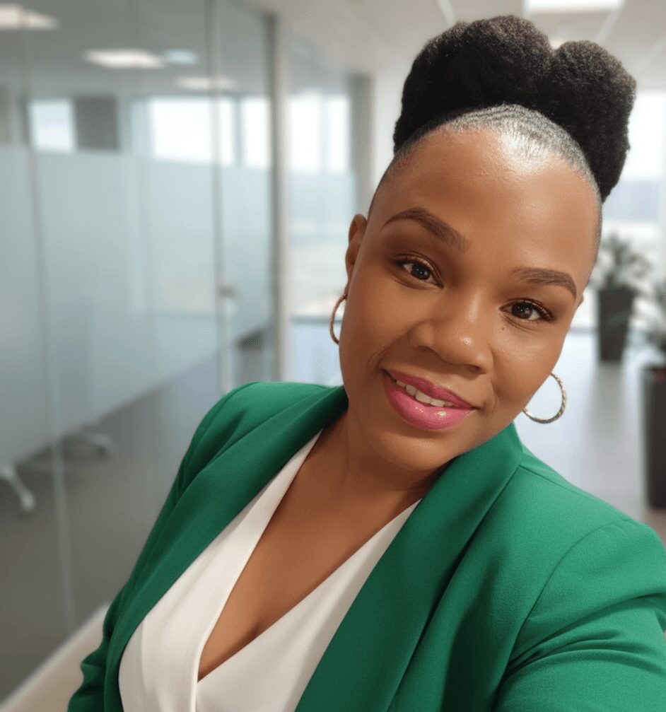 Woman with natural hair styled in a puff, wearing a green blazer and white top, smiling in an office setting.