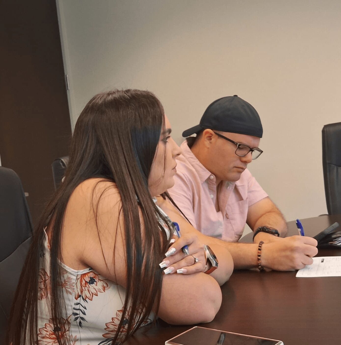 Two people sitting at a table, one with long hair and the other wearing glasses and a cap, writing on paper.