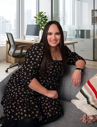 Woman sitting on a sofa in a modern office with large windows, a desk, and a computer in the background.