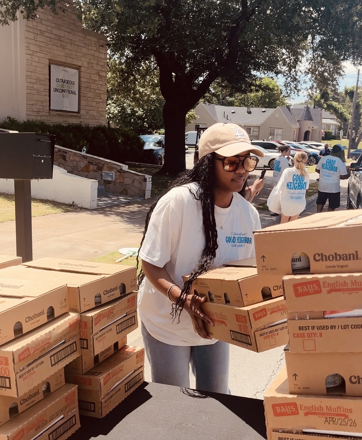 Woman with long hair, sunglasses, and a hat holding pizza boxes outdoors, with trees and people in the background.