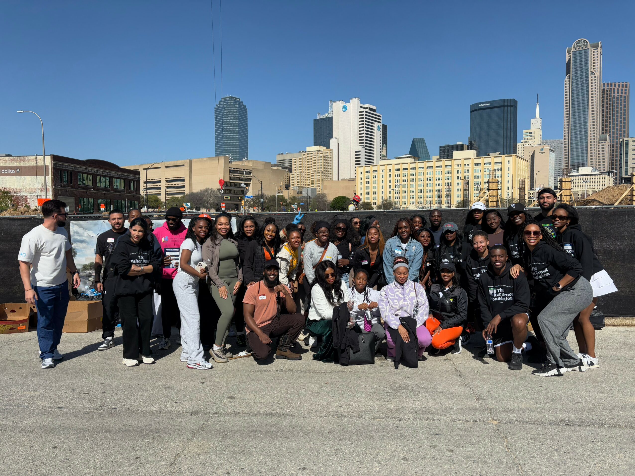 Group of people posing outdoors with city skyline in background, clear blue sky, some holding bags, diverse group, urban setting.
