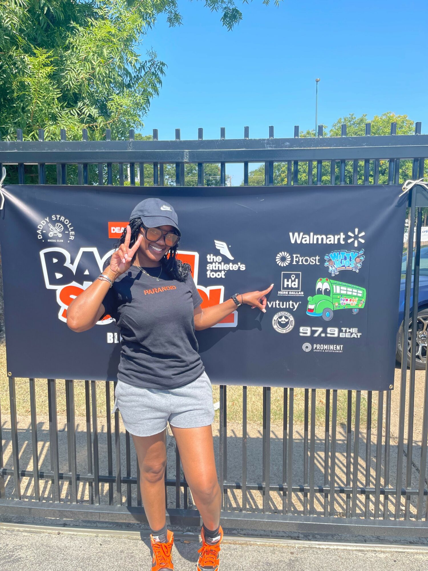 Person in gray shirt and shorts, wearing a cap, standing outdoors near a fence with a banner, making a peace sign.