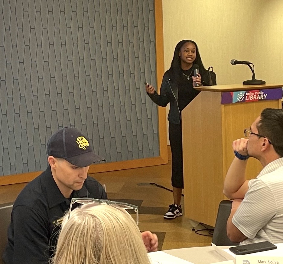 Young woman speaking at a podium in a library, with audience members listening attentively.