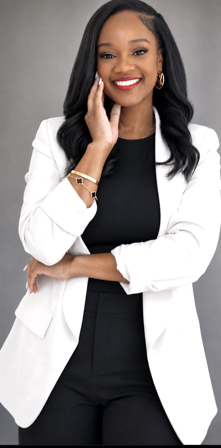 Woman with long black hair smiling, wearing a white blazer and black top, standing with arms crossed.