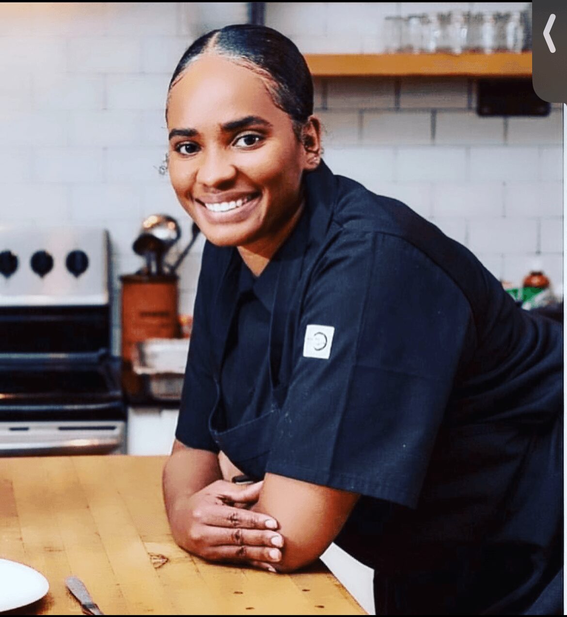 Person in a black uniform leaning on a wooden table, smiling, in a kitchen setting with white tiled wall.