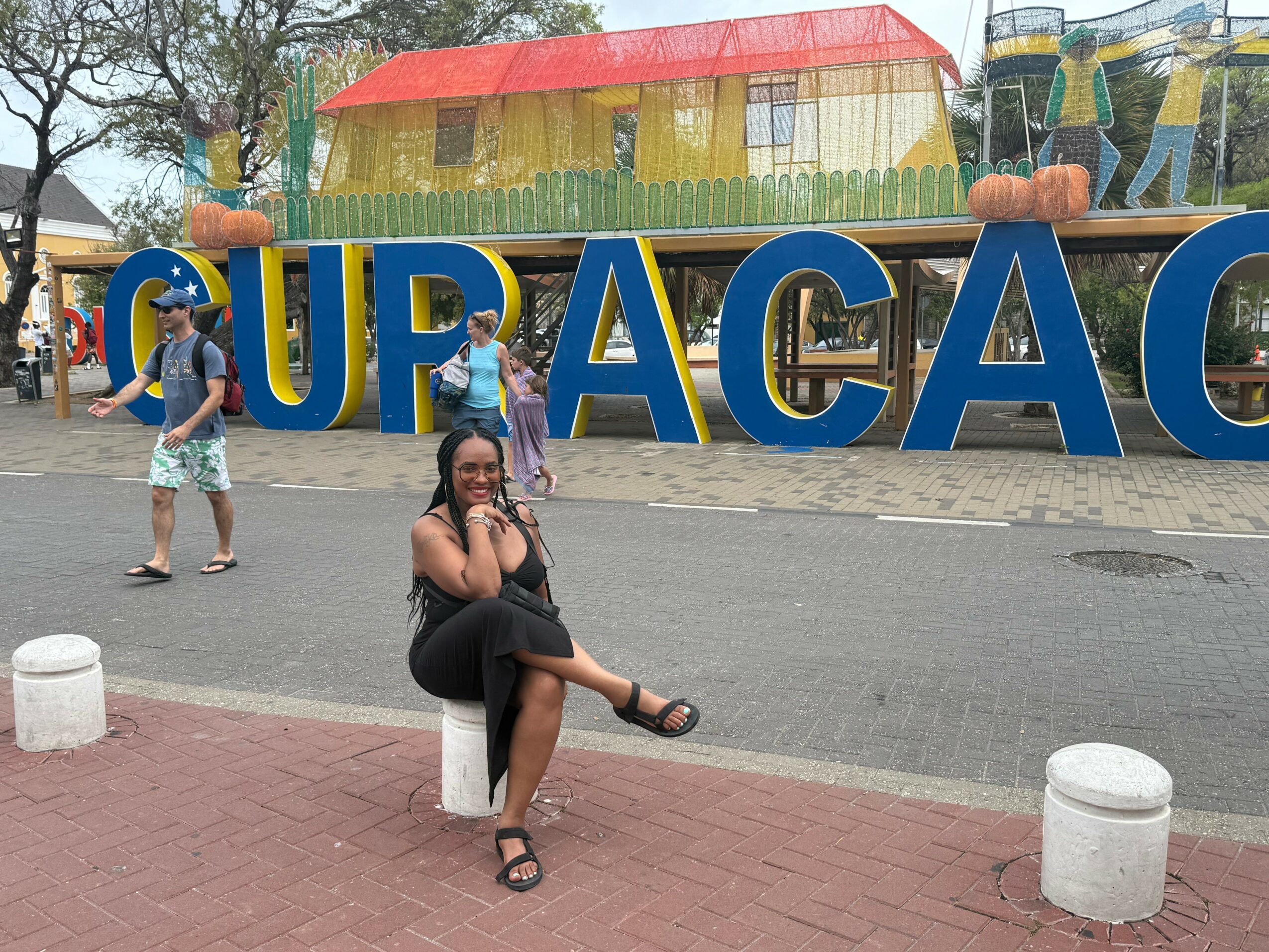 Woman sitting on a white bollard, smiling, with a colorful playground and large blue letters spelling 'CUPACAC' in the background.